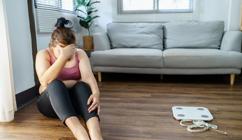 Hormonska neravnoteža: Frustrirana žena sedi na podu pored vage, simptom gojaznosti.